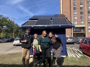 Four students pose in front of the solar shed in lot B behind Mortensen Hall. One is pointing to the solar panels on top of the shed.